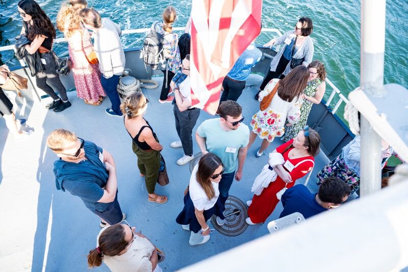 The image shows a group of people on the deck of a boat. They are dressed in casual clothes and appear to be enjoying themselves. Some are looking at the camera, while others are engaged in conversation. The boat is on the water, and the American flag is visible.